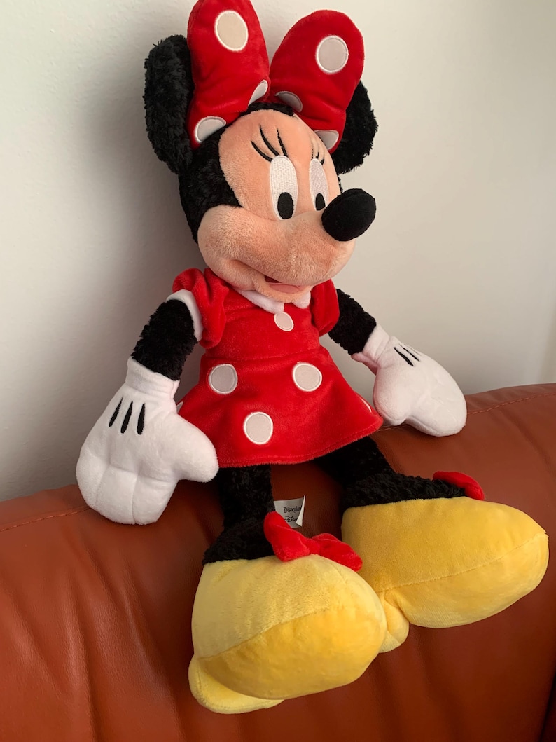 May include: A large plush toy of Minnie Mouse wearing a red dress with white polka dots and a red bow with white polka dots. She has black fur, a pink nose, and yellow shoes.
