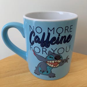 May include: A light blue ceramic mug with a cartoon Stitch character from Lilo & Stitch. The mug has the text "No More Caffeine For You" printed on it.