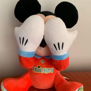 Puede incluir: Un juguete de peluche Mickey Mouse rojo y azul que lleva un pijama rojo y azul con la palabra "mickey" en la parte delantera. El juguete est&aacute; sentado con las manos cubri&eacute;ndose los ojos.