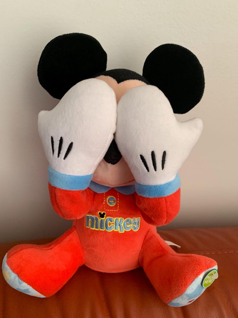 Puede incluir: Un juguete de peluche Mickey Mouse rojo y azul que lleva un pijama rojo y azul con la palabra "mickey" en la parte delantera. El juguete est&aacute; sentado con las manos cubri&eacute;ndose los ojos.
