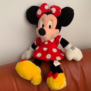 Może przedstawiać: Pluszowa zabawka przedstawiająca Minnie Mouse w czerwonej sukience w białe kropki i z czerwoną kokardką w białe kropki. Ma czarne uszy i ż&oacute;łtą twarz.