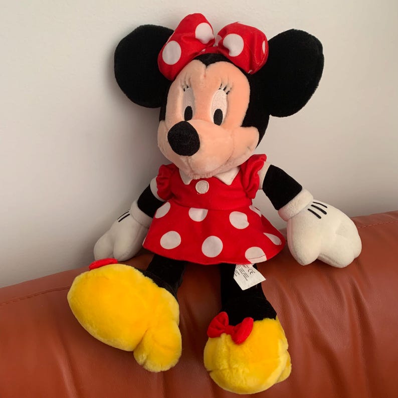 Może przedstawiać: Pluszowa zabawka przedstawiająca Minnie Mouse w czerwonej sukience w białe kropki i z czerwoną kokardką w białe kropki. Ma czarne uszy i ż&oacute;łtą twarz.