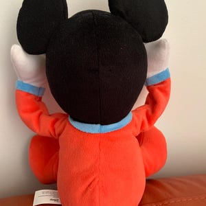 Puede incluir: Un juguete de peluche Mickey Mouse rojo y negro con un mono azul y blanco. El juguete est&aacute; sentado en una superficie marr&oacute;n. El juguete tiene un logotipo de Disney en la parte inferior.