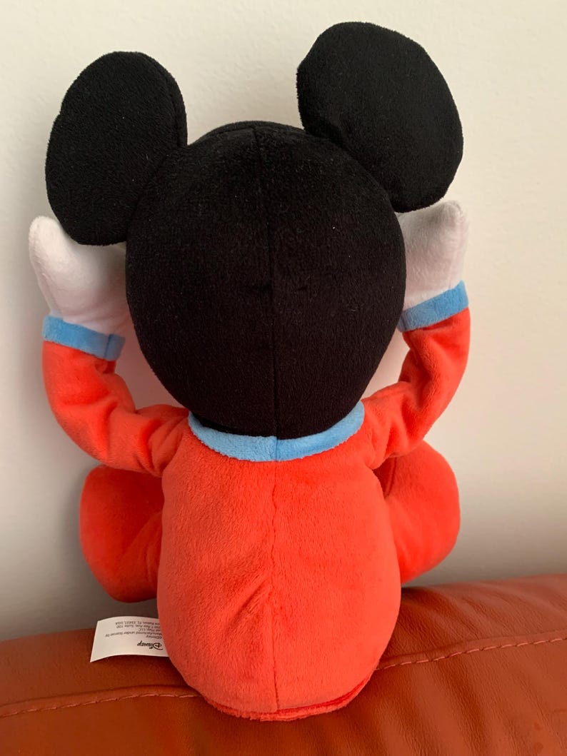 Puede incluir: Un juguete de peluche Mickey Mouse rojo y negro con un mono azul y blanco. El juguete est&aacute; sentado en una superficie marr&oacute;n. El juguete tiene un logotipo de Disney en la parte inferior.