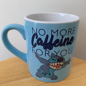 May include: A light blue ceramic mug with a cartoon Stitch character from Lilo & Stitch. The mug has the text "No More Caffeine For You" in black lettering.