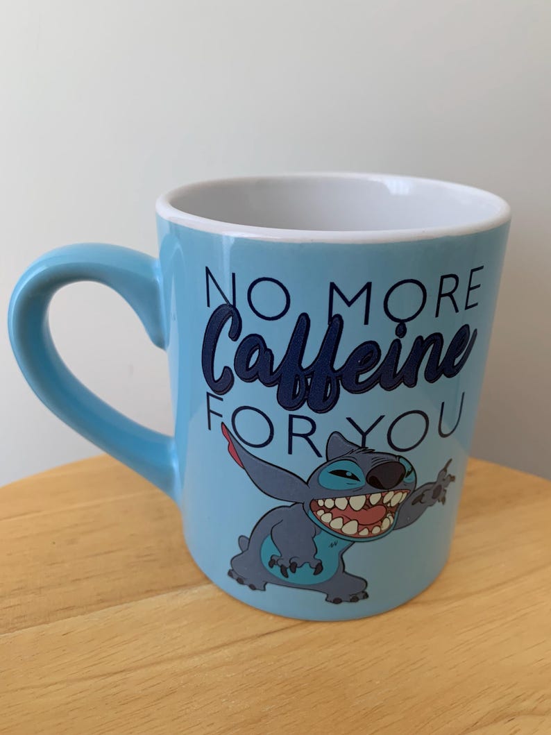 May include: A light blue ceramic mug with a cartoon Stitch character from Lilo & Stitch. The mug has the text "No More Caffeine For You" in black lettering.