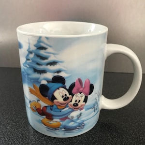 Peut inclure: Une tasse en céramique blanche avec une scène d'hiver représentant Mickey et Minnie Mouse faisant du patin à glace. L'arrière-plan est une forêt enneigée avec un pin bleu et blanc.