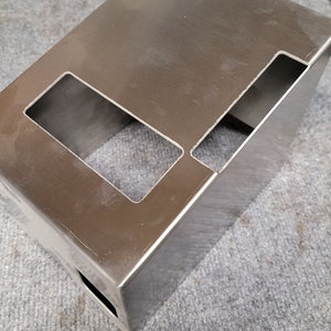 May include: A rectangular, brushed stainless steel box with cut-out rectangular openings on the top and side. The metal has a reflective surface and appears to be newly fabricated. The box is likely a component or housing for another product.