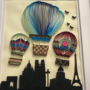 May include: A framed paper quilling art piece featuring three colorful hot air balloons above a black silhouette of a cityscape, including the Eiffel Tower. The balloons are blue, pink, and green with intricate paper coil details. The artwork is set against a cream-colored background.