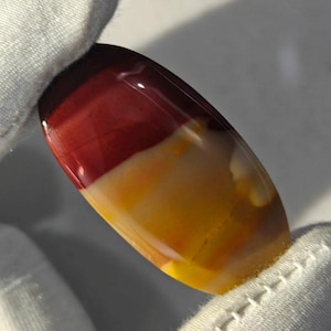 May include: A polished, oval-shaped cabochon gemstone featuring distinct horizontal bands of deep maroon, creamy white, and vibrant yellow. The stone has a smooth, reflective surface with a high-gloss finish, showcasing natural color transitions.