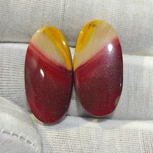 May include: Two oval-shaped cabochons featuring a polished finish with distinct color bands of deep red, cream, and bright yellow. The stones have a smooth, reflective surface and are displayed side-by-side, showcasing a natural layered pattern.