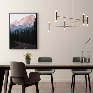 May include: A dining room with a modern light fixture hanging over a wooden table with six chairs. A framed photograph of a mountain range with a winding road is on the wall behind the table.