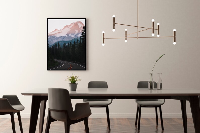 May include: A dining room with a modern light fixture hanging over a wooden table with six chairs. A framed photograph of a mountain range with a winding road is on the wall behind the table.