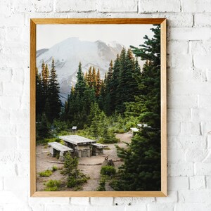 May include: A framed photograph of a mountain peak with snow-capped summit, surrounded by a lush forest of evergreen trees. A wooden picnic table sits in the clearing, with a dirt path leading to it.