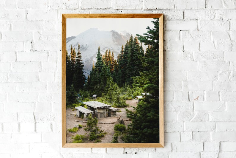 May include: A framed photograph of a mountain peak with snow-capped summit, surrounded by a lush forest of evergreen trees. A wooden picnic table sits in the clearing, with a dirt path leading to it.