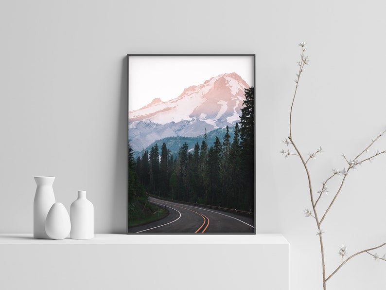 May include: A framed photograph of a winding road leading through a forest towards a snow-capped mountain peak. The mountain is a vibrant pink and blue color, with a clear sky above.