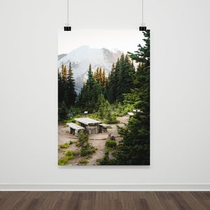 May include: A wooden picnic table sits in a clearing in a forest, surrounded by tall evergreen trees. A snow-capped mountain peak is visible in the background.
