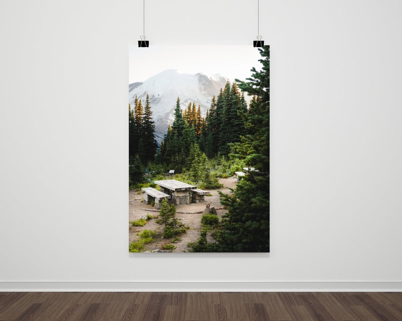 May include: A wooden picnic table sits in a clearing in a forest, surrounded by tall evergreen trees. A snow-capped mountain peak is visible in the background.