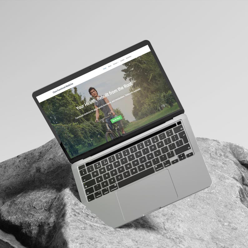K&ouml;nnte beinhalten: Ein silberner Laptop, der eine Website mit dem Text "Your Health, Rebuilt from the Root" anzeigt. Der Bildschirm zeigt eine Person, die in einem Park Fahrrad f&auml;hrt. Der Laptop liegt auf einem grauen, strukturierten Felsen.