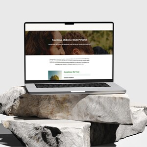 K&ouml;nnte beinhalten: Ein silberner Laptop, der eine Website mit dem Text "Functional Medicine, Made Personal" und einem Foto von zwei Personen anzeigt. Der Laptop steht auf einem Stapel gro&szlig;er, hellgrauer Steine. Der Inhalt der Website enth&auml;lt Informationen &uuml;ber funktionelle Medizin.