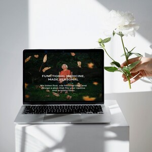 Puede incluir: Un port&aacute;til plateado que muestra el texto "FUNCTIONAL MEDICINE, MADE PERSONAL" con una mujer con una camisa naranja en la pantalla. Una flor blanca se sostiene cerca del port&aacute;til. El port&aacute;til est&aacute; sobre una superficie blanca.