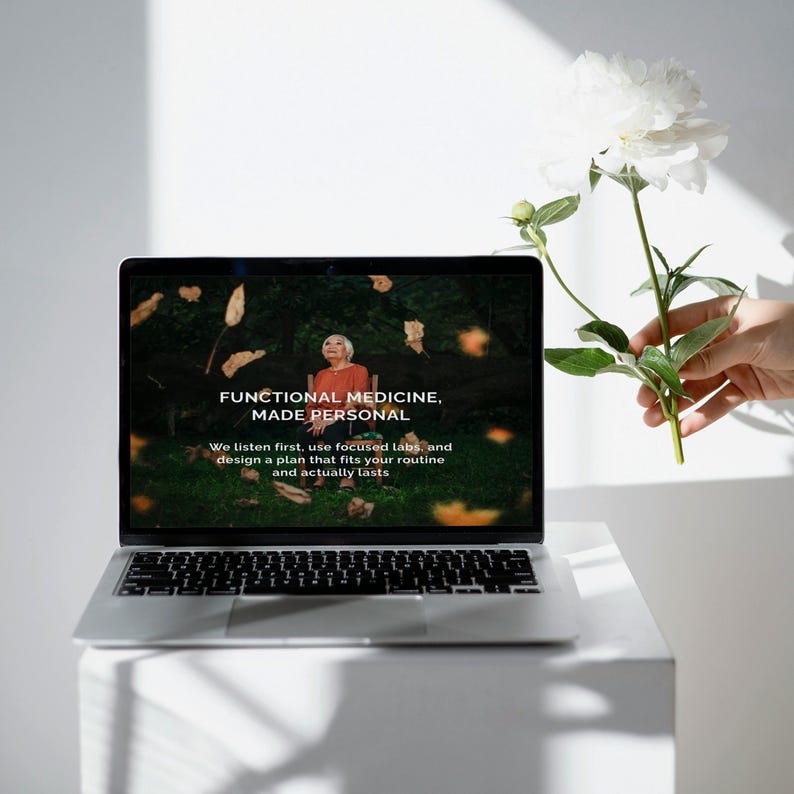 Puede incluir: Un port&aacute;til plateado que muestra el texto "FUNCTIONAL MEDICINE, MADE PERSONAL" con una mujer con una camisa naranja en la pantalla. Una flor blanca se sostiene cerca del port&aacute;til. El port&aacute;til est&aacute; sobre una superficie blanca.