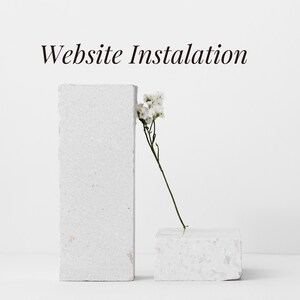 以下が含まれることがあります： 「Website Installation」のテキストが上にあり、白く質感のある長方形のブロックが2つ配置されたミニマリストな構図。小さなブロックには白い花の繊細な茎が寄りかかっており、穏やかでクリーンな美しさを演出しています。