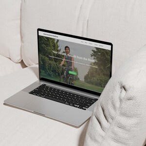 K&ouml;nnte beinhalten: Ein silberner Laptop, der eine Website mit dem Text "Your Health, Rebuilt from the Root" anzeigt. Der Bildschirm zeigt eine Person, die in einem Park Fahrrad f&auml;hrt. Der Laptop ist auf einer wei&szlig;en Oberfl&auml;che ge&ouml;ffnet, mit einer schwarzen Tastatur und einem schwarzen Bildschirmrand.