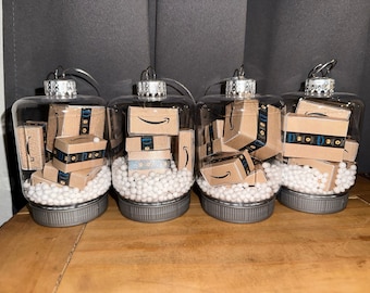 May include: Four clear glass ornaments filled with white packing peanuts and miniature cardboard boxes with the Amazon logo. The ornaments are decorated with silver tops and are ready to be hung on a Christmas tree.