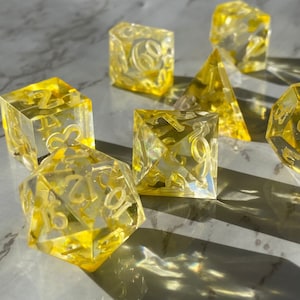 May include: A set of seven yellow transparent dice with white numbers. The dice are made of a clear material and have a frosted finish. The numbers on the dice are visible through the clear material.