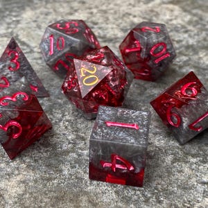 Puede incluir: Conjunto de siete dados poliédricos con un diseño de mármol negro y rojo. Los dados están numerados del 1 al 20 y tienen detalles dorados.
