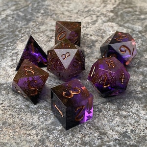 Könnte beinhalten: Satz von sieben lila und goldenen Würfeln mit glitzernder Oberfläche. Die Würfel sind von 1 bis 6 nummeriert, mit einem Unendlichkeitszeichen auf dem d8.