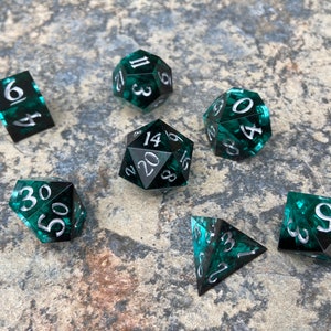 Op de afbeelding: Een set van zeven polyhedrale dobbelstenen in verschillende vormen, waaronder een d4, d6, d8, d10, d12, d20 en een percentagedobbelsteen. De dobbelstenen zijn doorschijnend teal met zilveren cijfers.