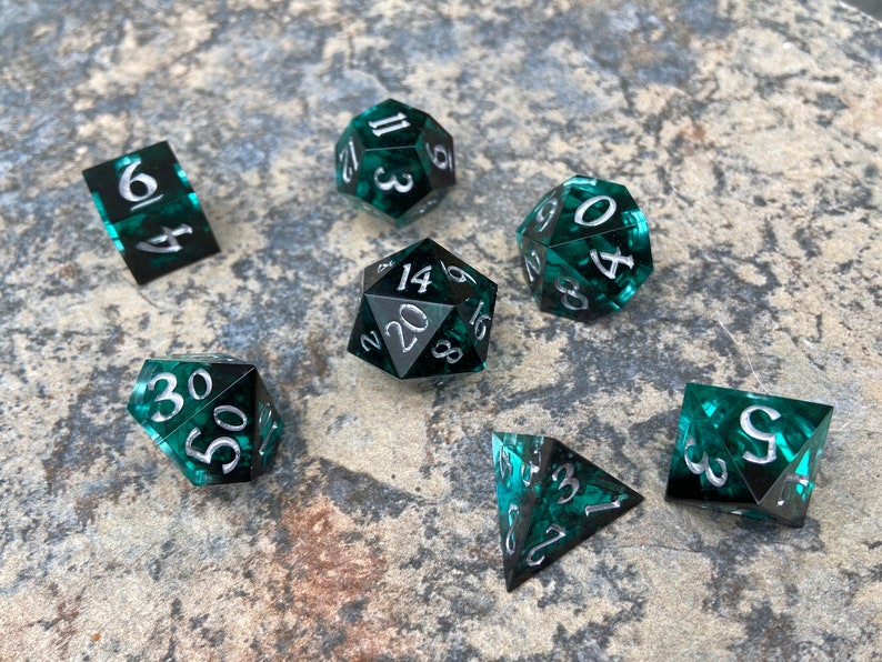 Op de afbeelding: Een set van zeven polyhedrale dobbelstenen in verschillende vormen, waaronder een d4, d6, d8, d10, d12, d20 en een percentagedobbelsteen. De dobbelstenen zijn doorschijnend teal met zilveren cijfers.