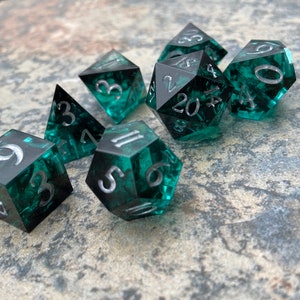 Op de afbeelding: Set van zeven polyhedrale dobbelstenen met een teal en zwart gemarmerd effect. Elke dobbelsteen heeft zilveren cijfers. De dobbelstenen hebben verschillende vormen, waaronder d4, d6, d8, d10, d12 en d20, voor rollenspellen.