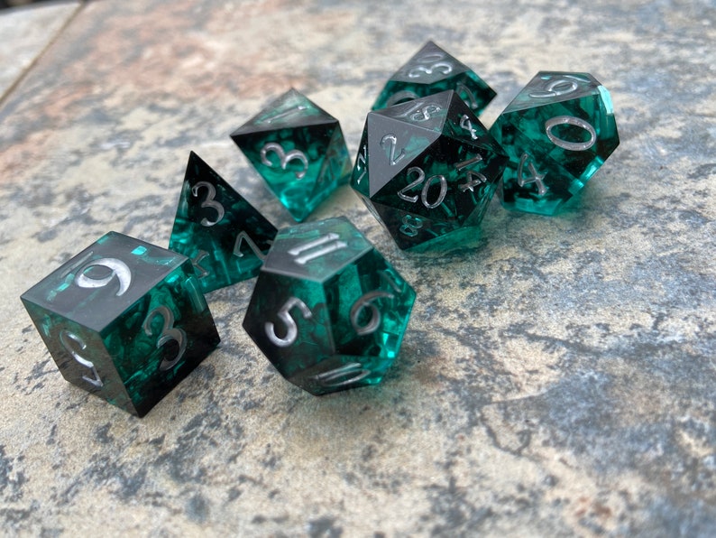 Op de afbeelding: Set van zeven polyhedrale dobbelstenen met een teal en zwart gemarmerd effect. Elke dobbelsteen heeft zilveren cijfers. De dobbelstenen hebben verschillende vormen, waaronder d4, d6, d8, d10, d12 en d20, voor rollenspellen.