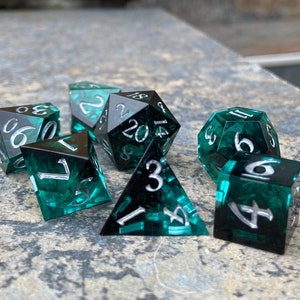 Op de afbeelding: Een set van zeven polyhedrale dobbelstenen voor tabletopspellen. De dobbelstenen zijn doorschijnend teal met een wervelpatroon en zilveren cijfers. De set bevat een d4, d6, d8, d10, d12, d20 en een percentagedobbelsteen.