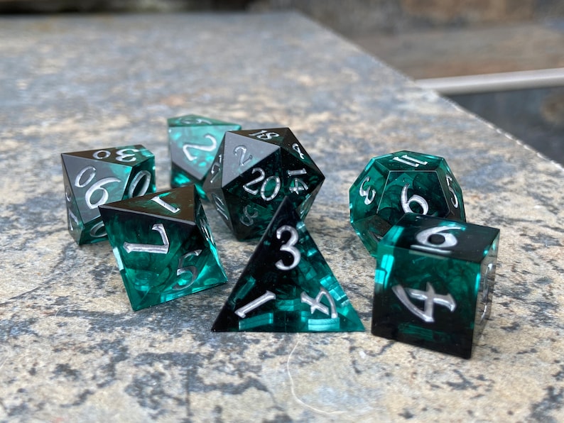 Op de afbeelding: Een set van zeven polyhedrale dobbelstenen voor tabletopspellen. De dobbelstenen zijn doorschijnend teal met een wervelpatroon en zilveren cijfers. De set bevat een d4, d6, d8, d10, d12, d20 en een percentagedobbelsteen.