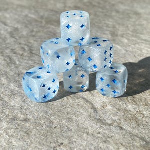Pode incluir: Um conjunto de seis dados transparentes com glitter azul e marcas em forma de estrela azul.
