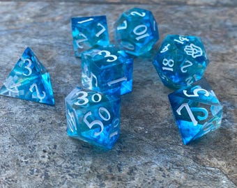 May include: A set of seven blue and white dice with white numbers. The dice are made of clear plastic with a swirling blue pattern inside. The dice are a d4, d6, d8, d10, d%, d12, and d20.
