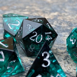 Op de afbeelding: Set polyhedrale dobbelstenen voor tabletopspellen. De dobbelstenen zijn diep teal van kleur met zilveren cijfers. De d20 dobbelsteen staat in het midden, omringd door andere dobbelstenen. De dobbelstenen hebben een gemarmerd effect.
