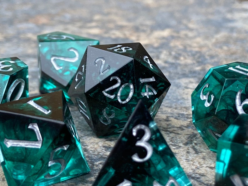 Op de afbeelding: Set polyhedrale dobbelstenen voor tabletopspellen. De dobbelstenen zijn diep teal van kleur met zilveren cijfers. De d20 dobbelsteen staat in het midden, omringd door andere dobbelstenen. De dobbelstenen hebben een gemarmerd effect.
