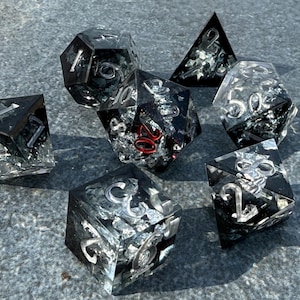 Puede incluir: Conjunto de siete dados negros y plateados con resina transparente y purpurina plateada. Los dados están numerados del 1 al 20.