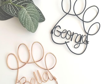 May include: Two wire art pieces shaped like animal paw prints. One is made of black wire with the name George formed in cursive script, and the other is made of copper-colored wire with the name Bella formed in cursive script.