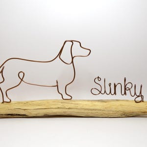 May include: Wire sculpture of a dachshund dog standing on a piece of driftwood. The dog's name, "Slinky", is written in wire below the dog.