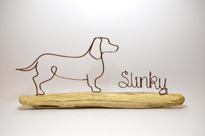 May include: Wire sculpture of a dachshund dog standing on a piece of driftwood. The dog's name, "Slinky", is written in wire below the dog.