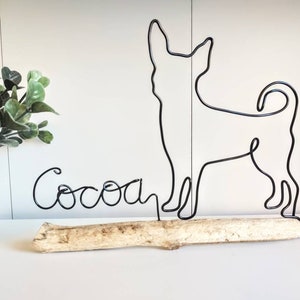 May include: Black wire sculpture of a chihuahua dog standing on a piece of driftwood with the name "Cocoa" written in wire below it.