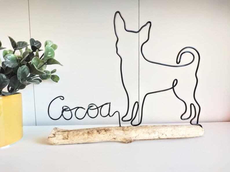 May include: Black wire sculpture of a chihuahua dog standing on a piece of driftwood with the name "Cocoa" written in wire below it.