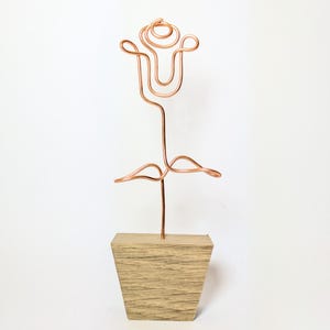 May include: A copper wire sculpture shaped like a rose flower with a single stem and two leaves, mounted into a light brown, trapezoidal wooden base. The metallic wire is bent into a continuous outline to form the petals and foliage.
