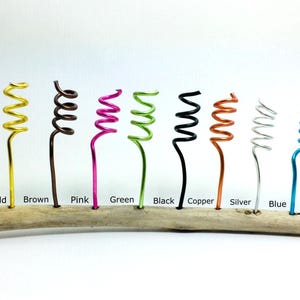May include: A piece of driftwood with ten coiled wire stems in different colors: gold, brown, pink, green, black, copper, silver, blue, turquoise, and purple.